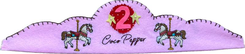 Lilac felt crown with two embroidered carousel horses, the name Coco Pepper in script, and a pink number 2 on a red polka-dot patch with yellow stars.