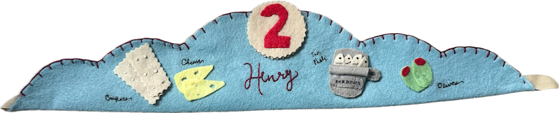 Pale-blue felt crown laid out like a snack plate: a felt cracker, a wedge of cheese, a tin of sardines and two olives, each labeled in tiny stitching, with the name Henry and a red number 2.