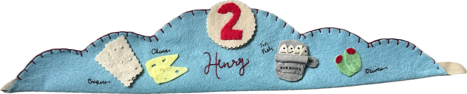 Pale-blue felt crown laid out like a snack plate: a felt cracker, a wedge of cheese, a tin of sardines and two olives, each labeled in tiny stitching, with the name Henry and a red number 2.