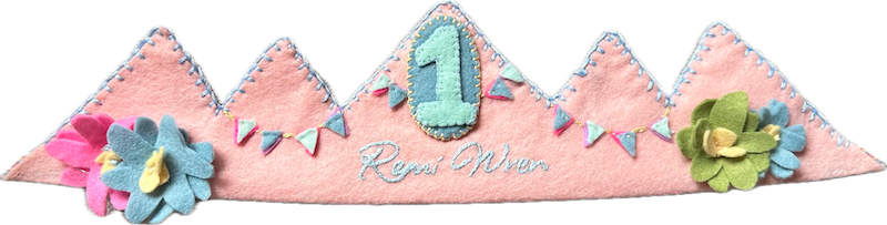 Blush-pink pointed felt crown with tiny felt bunting, layered felt flowers in pink, blue and green, the name Remi Wren in white script, and a mint number 1 patch, tied with rainbow ribbon.