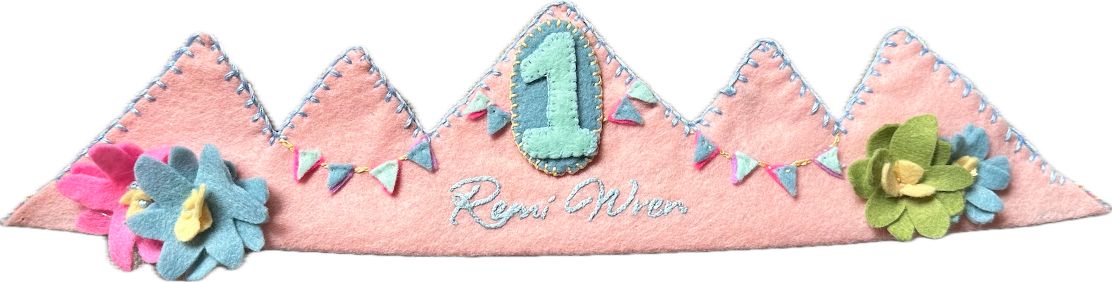 Blush-pink pointed felt crown with tiny felt bunting, layered felt flowers in pink, blue and green, the name Remi Wren in white script, and a mint number 1 patch, tied with rainbow ribbon.